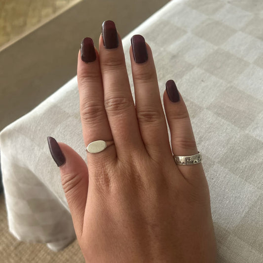 A close-up of a hand with dark plum nail polish, wearing a simple oval white ring on one finger and a textured silver band on the other, resting on a white checkered tablecloth.