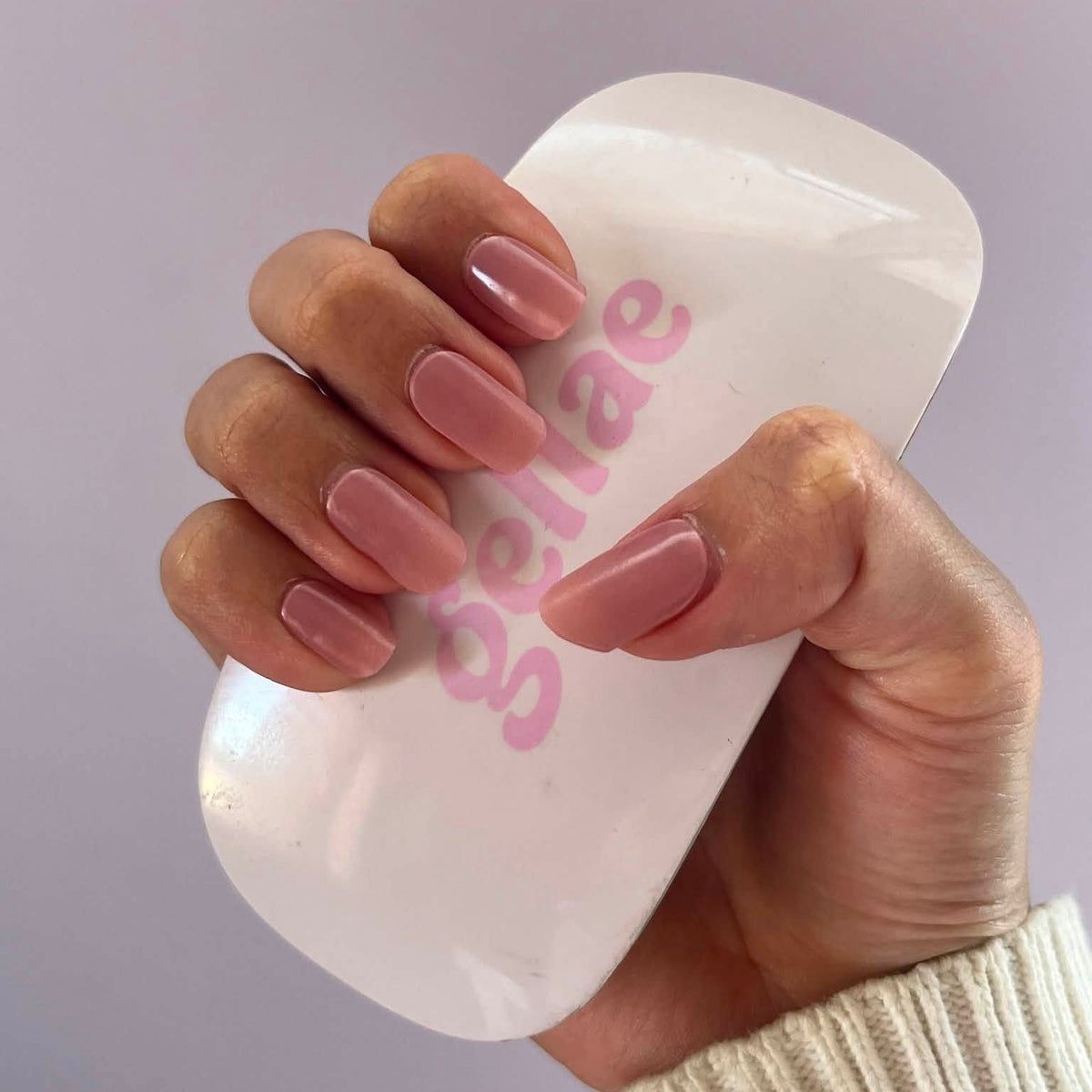 A hand with a glossy mauve-pink manicure holds a white curved nail file or press tool with pink 'schae' branding against a soft lavender background.
