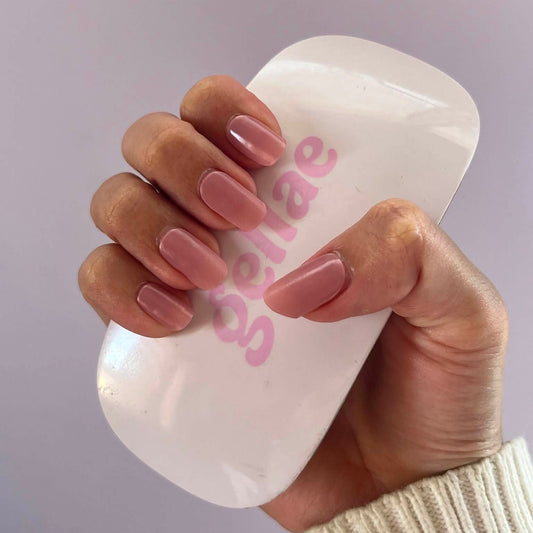 A hand with a glossy mauve-pink manicure holds a white curved nail file or press tool with pink 'schae' branding against a soft lavender background.