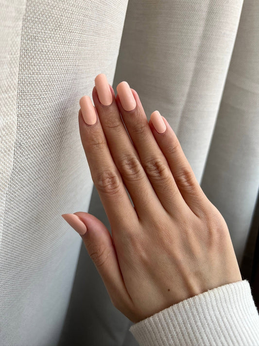 A close-up of a hand with a matte nude pink manicure resting against a textured light gray curtain.